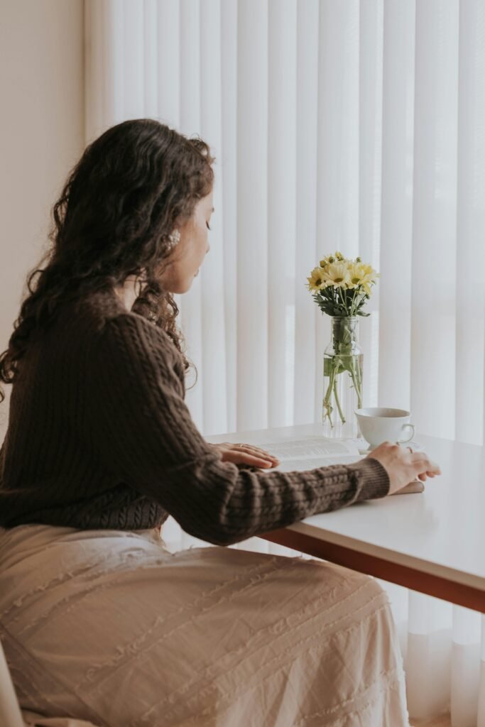Thoughtful woman sitting indoors by flowers and coffee, bathed in soft daylight.