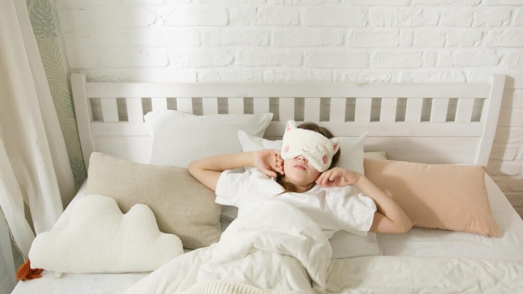 A young girl resting in bed with a cute sleep mask surrounded by cozy pillows.